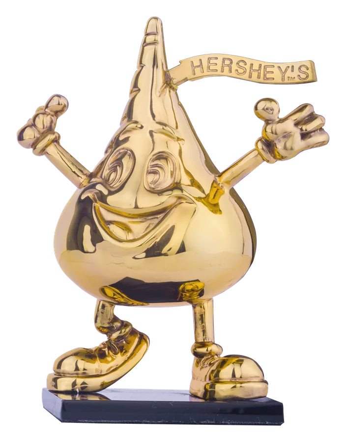 Den Hershey’s-Kiss-Charakter mat opgehuewenen Äerm, a poléiertem 24-Karat-Gold, mam Hershey’s-Banner op der Spëtzt, op engem schwaarze Sockel
